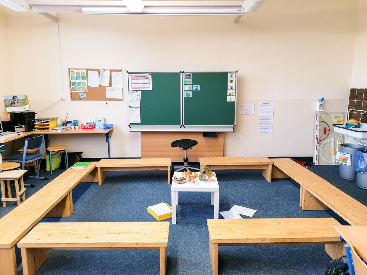 Klassenraum mit Bänken und kleinem weißen Tisch vor der Schultafel in der Grundschule der DPFA-Regenbogen-Schulen Rabenau.